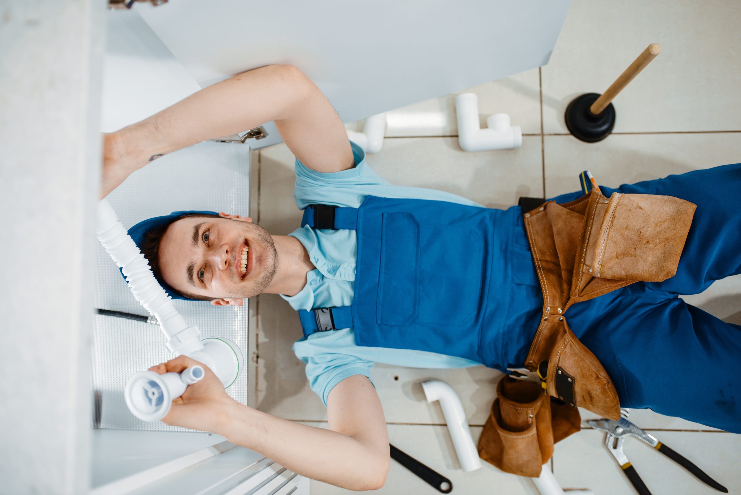 Plombier masculin en uniforme installant un tuyau de vidange dans la cuisine, vue de dessus. Femme à tout faire avec évier de réparation de sac à outils, service d'équipement sanitaire à la maison